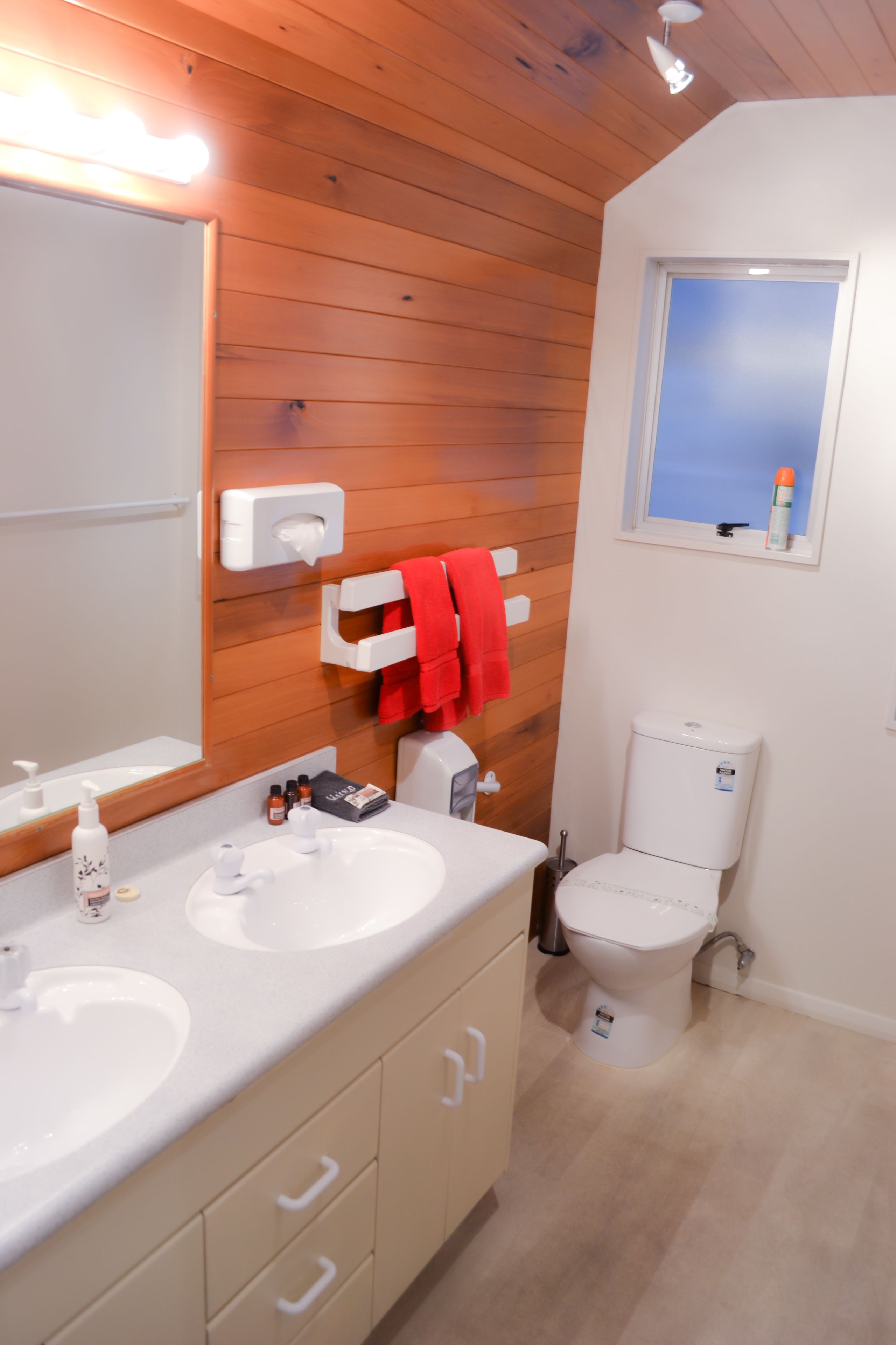 Un bagno moderno con rivestimenti in legno, uno specchio grande, due lavandini, un water e una finestra con vista.
