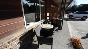 Un espacio al aire libre con sillas de mimbre y una mesa redonda, una pared de ladrillo y un ventanal que da a un estacionamiento.