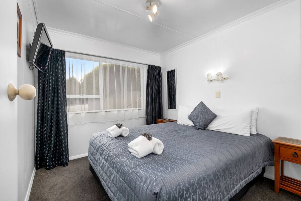 Ein Hotelzimmer mit einem großen Bett, einem Nachttisch, einem Wandfernseher und einem Fenster mit Vorhängen.
