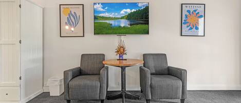 Een kamer met twee grijze fauteuils, een ronde houten tafel en drie ingelijste kunstwerken aan de muur.