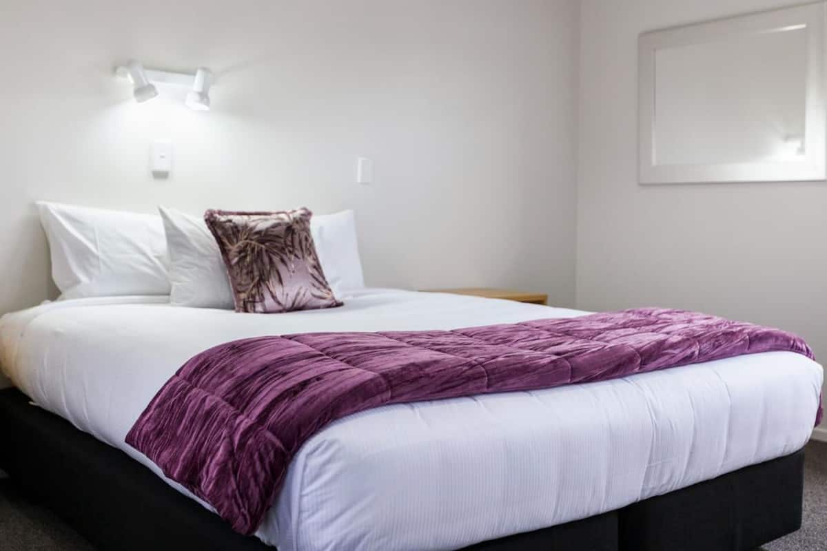 A neatly made bed with white and purple bedding, a single decorative pillow, and a wall-mounted light fixture.