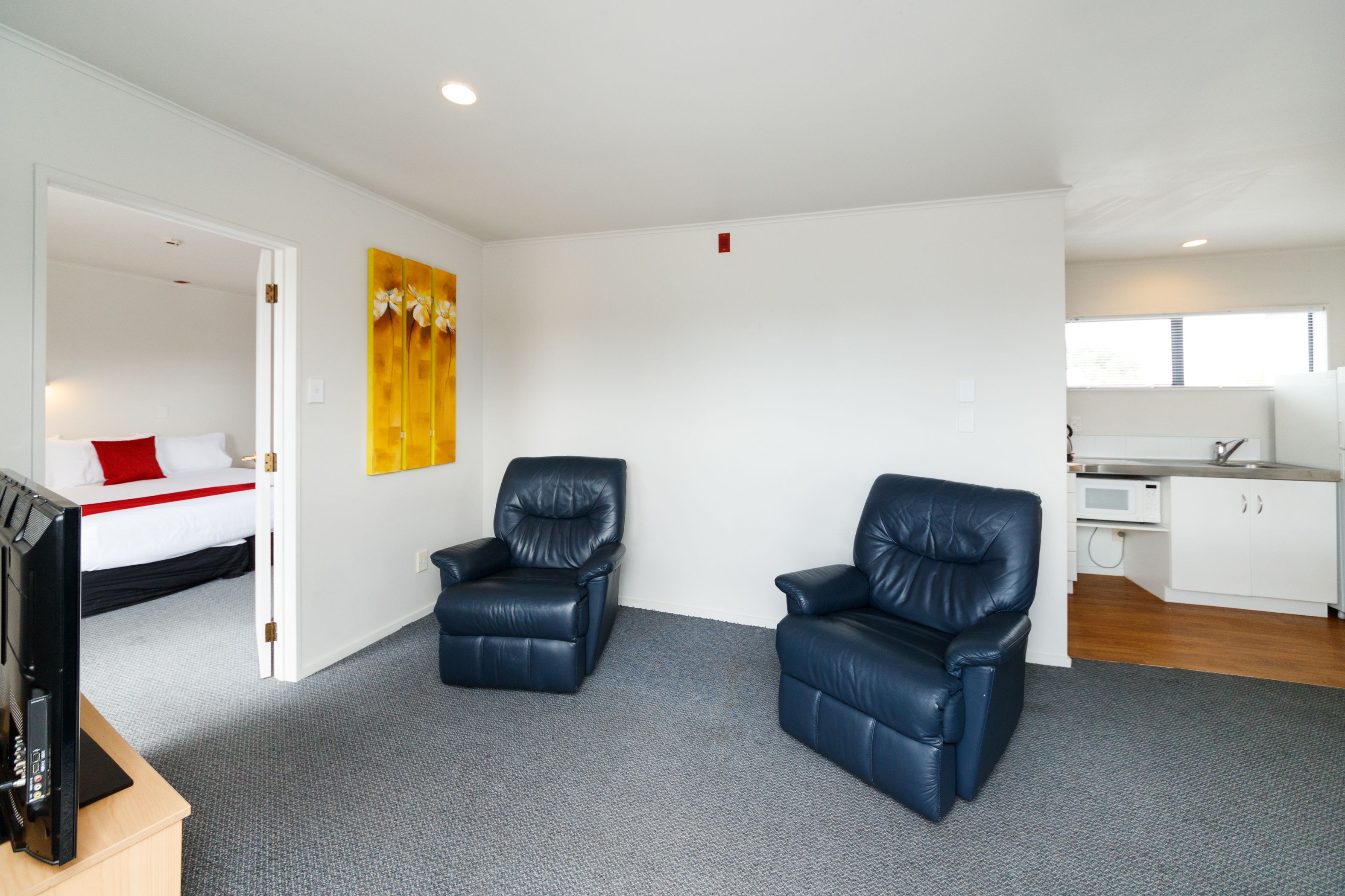 A modern hotel room with a grey carpet, two black leather chairs, a bed with white and red bedding, a yellow artwork on the wall, and a kitchenette in the background.