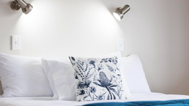 A bed with white bedding and a blue floral pillow, two wall-mounted lights, and a white wall with a light switch. - Drovers Motor Inn (Palmerston North)