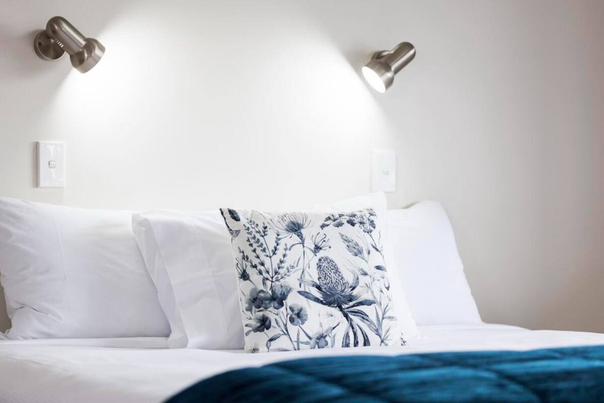 A bed with white bedding and a blue floral pillow, two wall-mounted lights, and a white wall with a light switch.