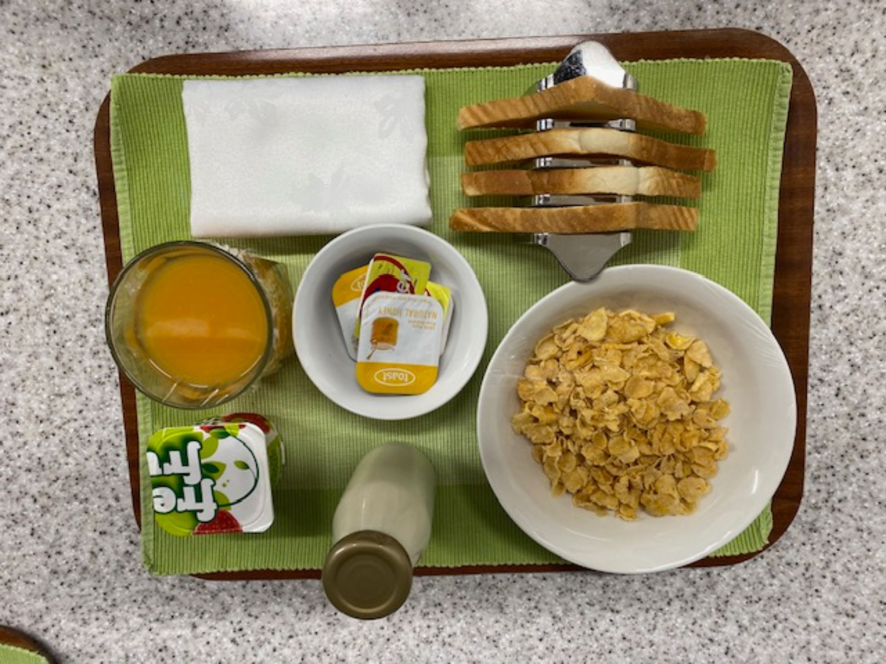 A breakfast tray with cereal, juice, a bottle of milk, a packet of butter, and a slice of bread.