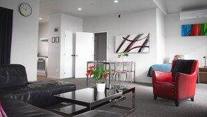 A modern living room with a sofa, a red chair, a coffee table with a vase of flowers, a dining area with chairs, a kitchen in the background, and a large abstract painting on the wall. - Drovers Motor Inn (Palmerston North)