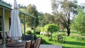 A patio with a wooden table and chairs, a white umbrella, and a view of a lush garden with a fence and trees. - Halls on Falls Homestead (Strath Creek)