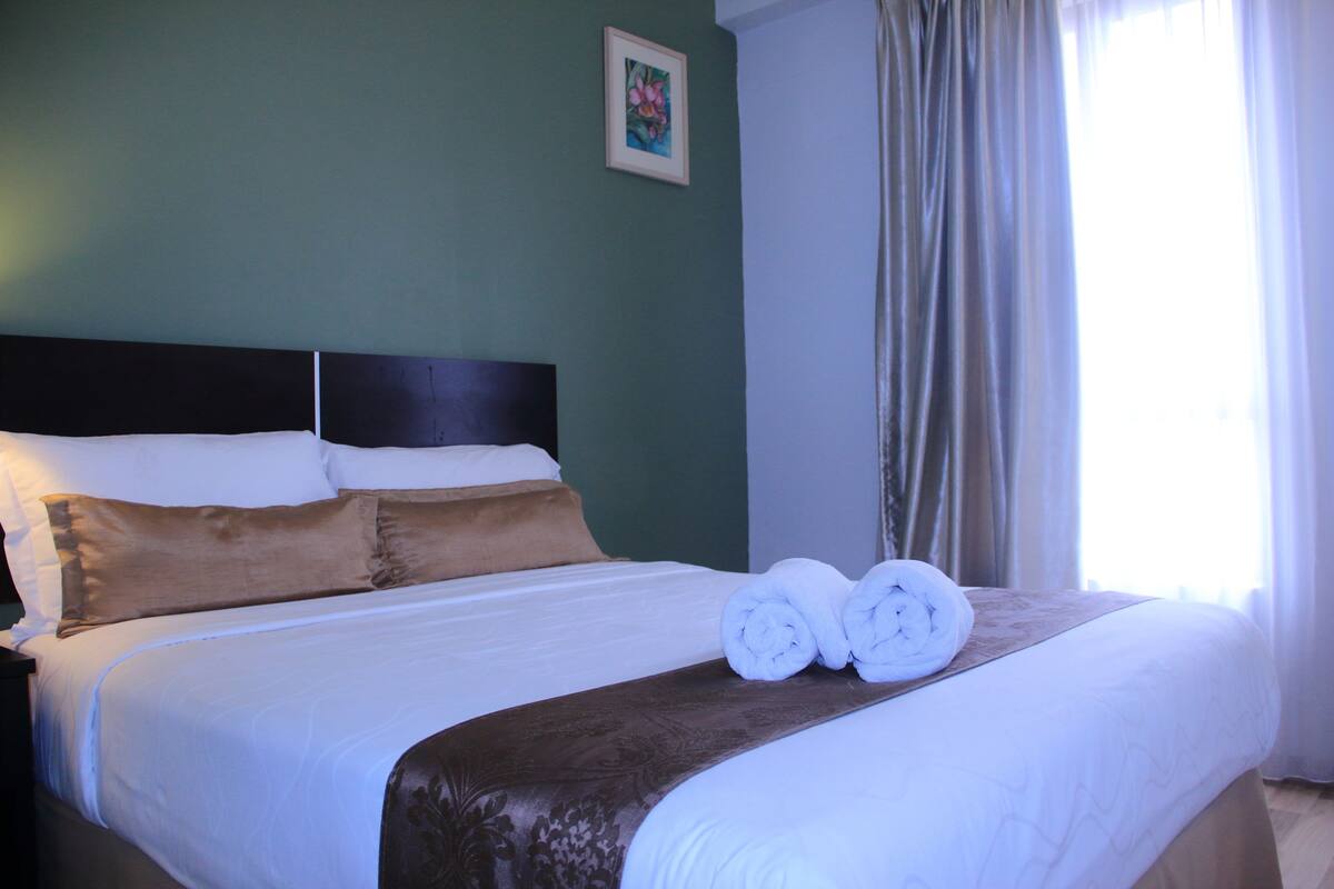 A neatly made bed with white and brown linens, a headboard, and two rolled towels on the bed.