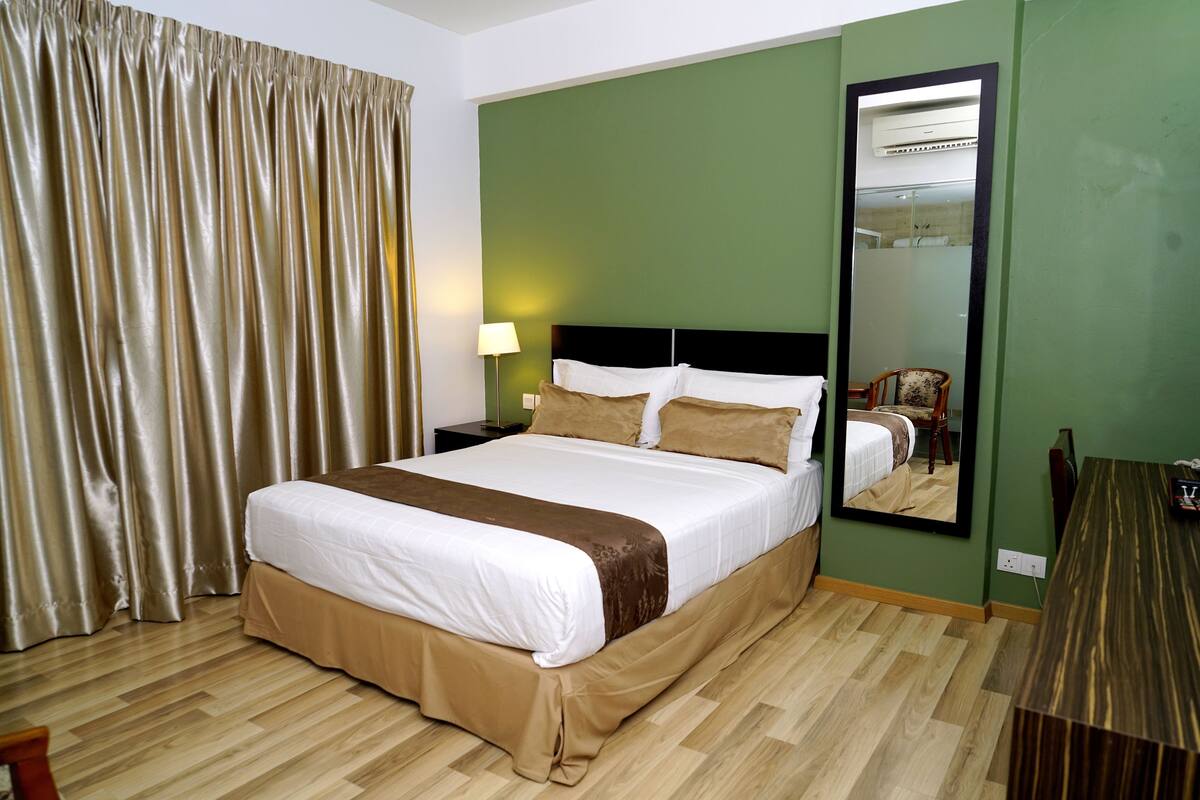 A hotel room with a large bed, a nightstand, a lamp, a mirror, and a wooden desk.