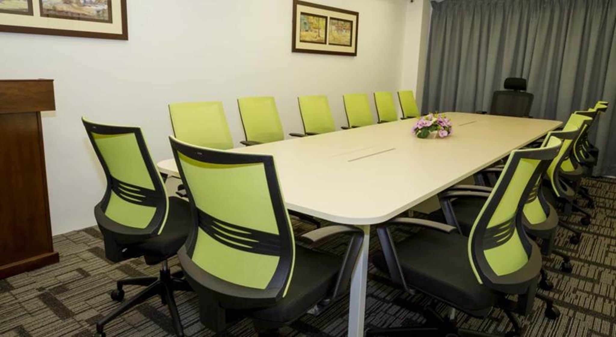 A conference room with a long table and green chairs, a podium, and framed artwork on the walls.