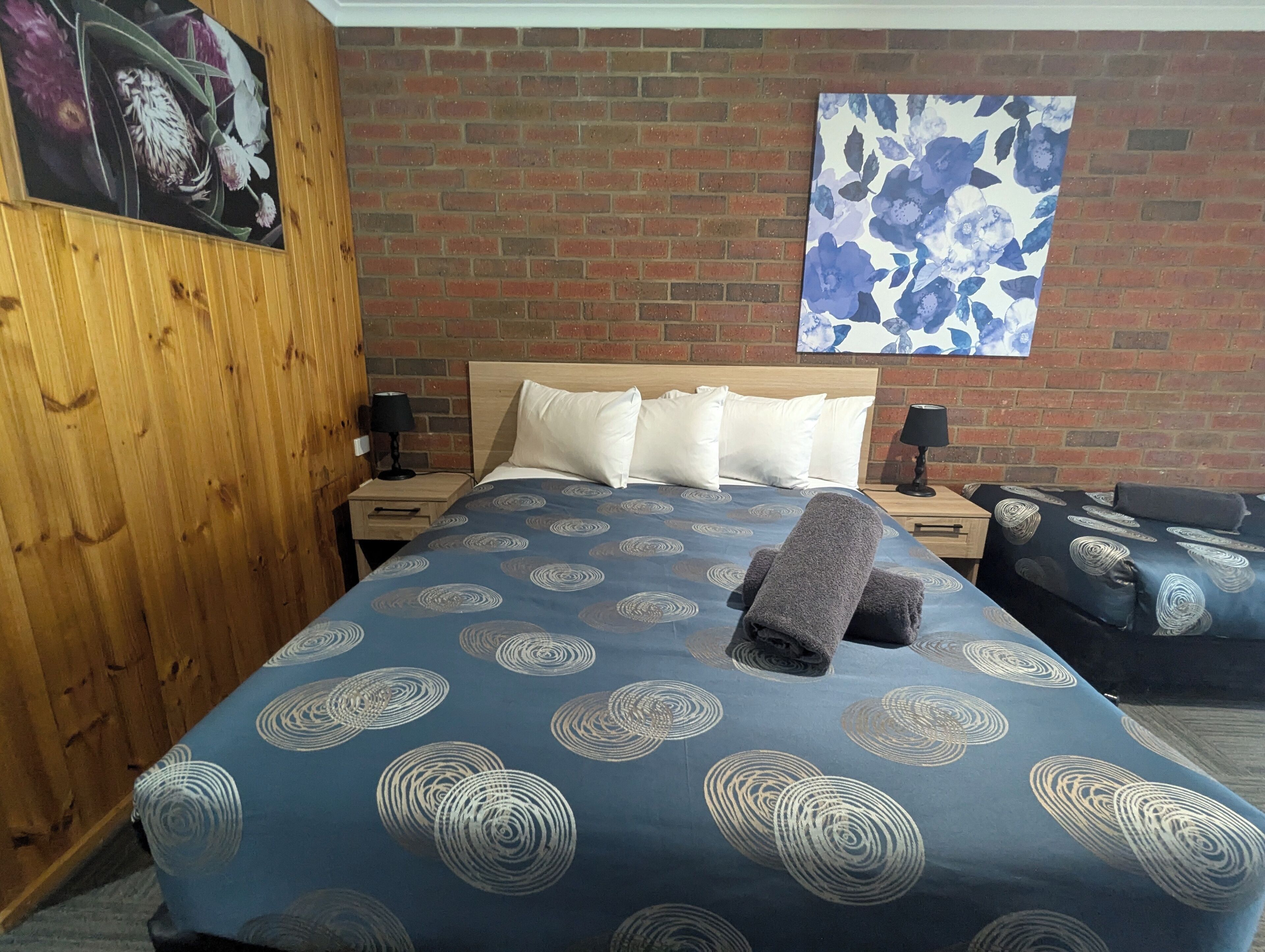 A hotel room with a brick wall, a wooden wardrobe, a bed with a blue patterned bedspread, white pillows, and a blue floral artwork on the wall.