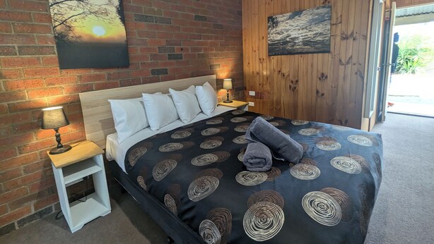 A bedroom with a brick wall, a wooden panel wall, a bed with a patterned bedspread, two bedside tables with lamps, and artwork on the walls.