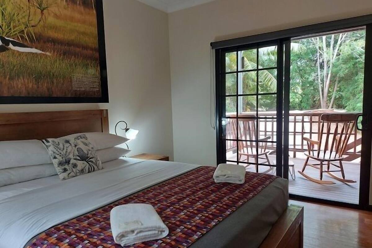 A hotel room with a bed, a nightstand, a lamp, and a view of an outdoor deck with chairs and a table.