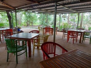 An outdoor covered dining area with multiple tables and chairs, surrounded by lush greenery.