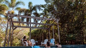 Een zwembadgedeelte met een houten pergola, witte ligbedden en mensen die ontspannen bij het zwembad.