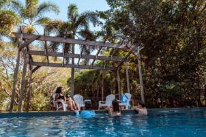 A pool area with a wooden pergola, white lounge chairs, and people relaxing by the pool.