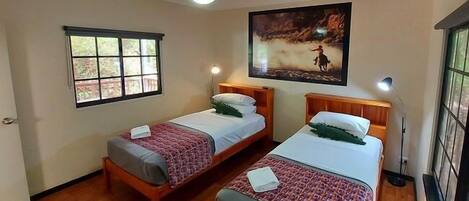 A room with two wooden beds, a large window, a ceiling fan, and a painting on the wall.