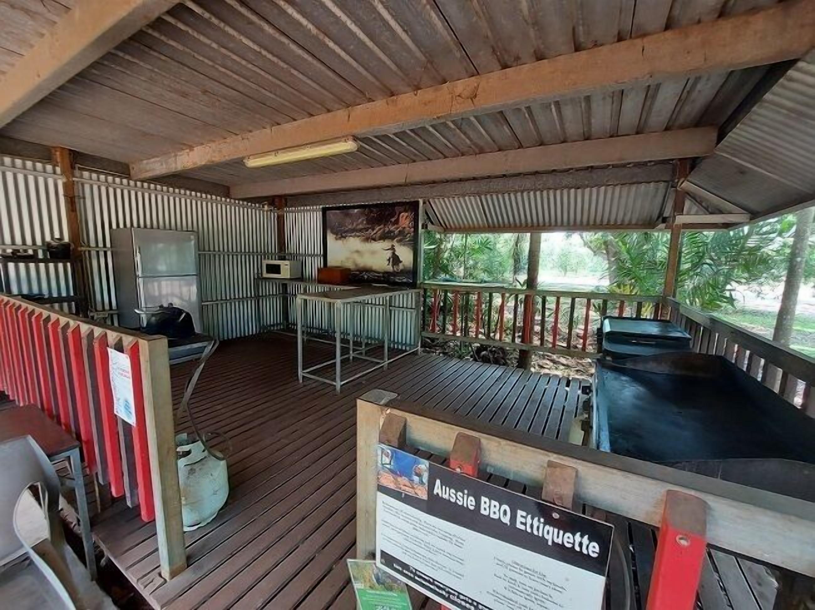 Zona de barbacoa al aire libre con terraza de madera, parrilla metálica y un cartel con normas para la barbacoa.
