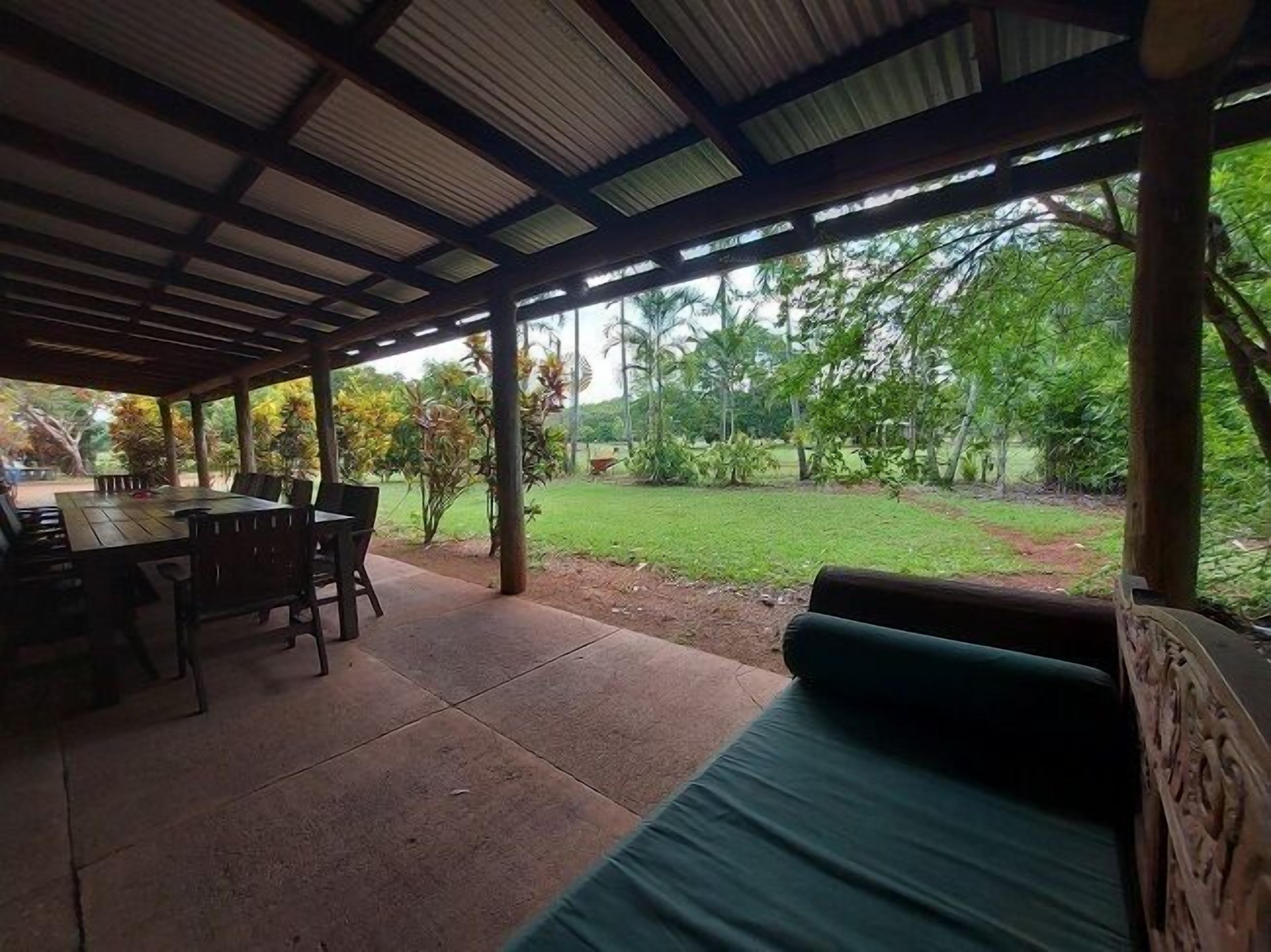 A covered outdoor area with a table and chairs, a cushioned bench, and a view of a grassy area with trees.