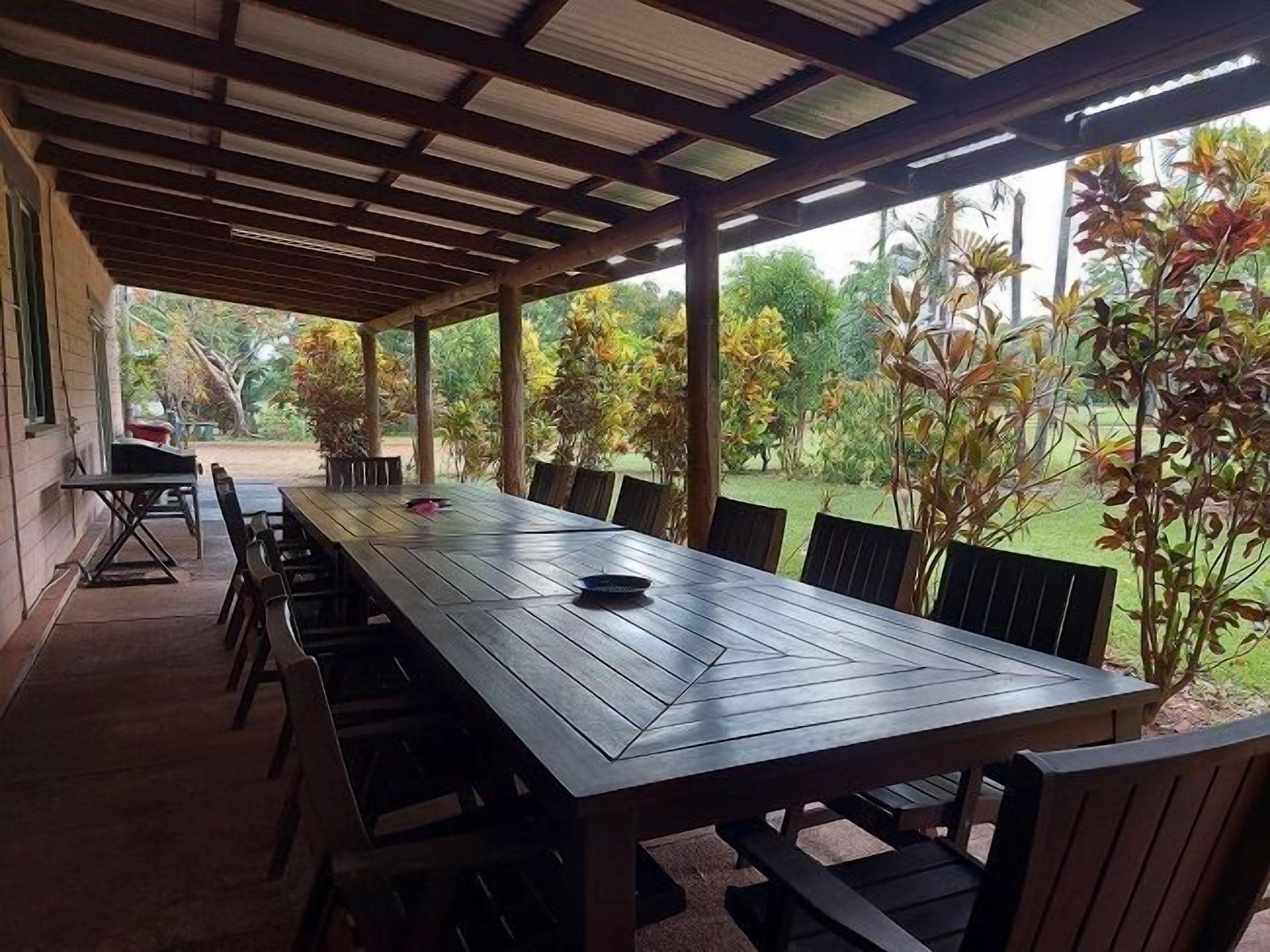 An outdoor covered patio with a long wooden table and chairs, surrounded by greenery and trees.