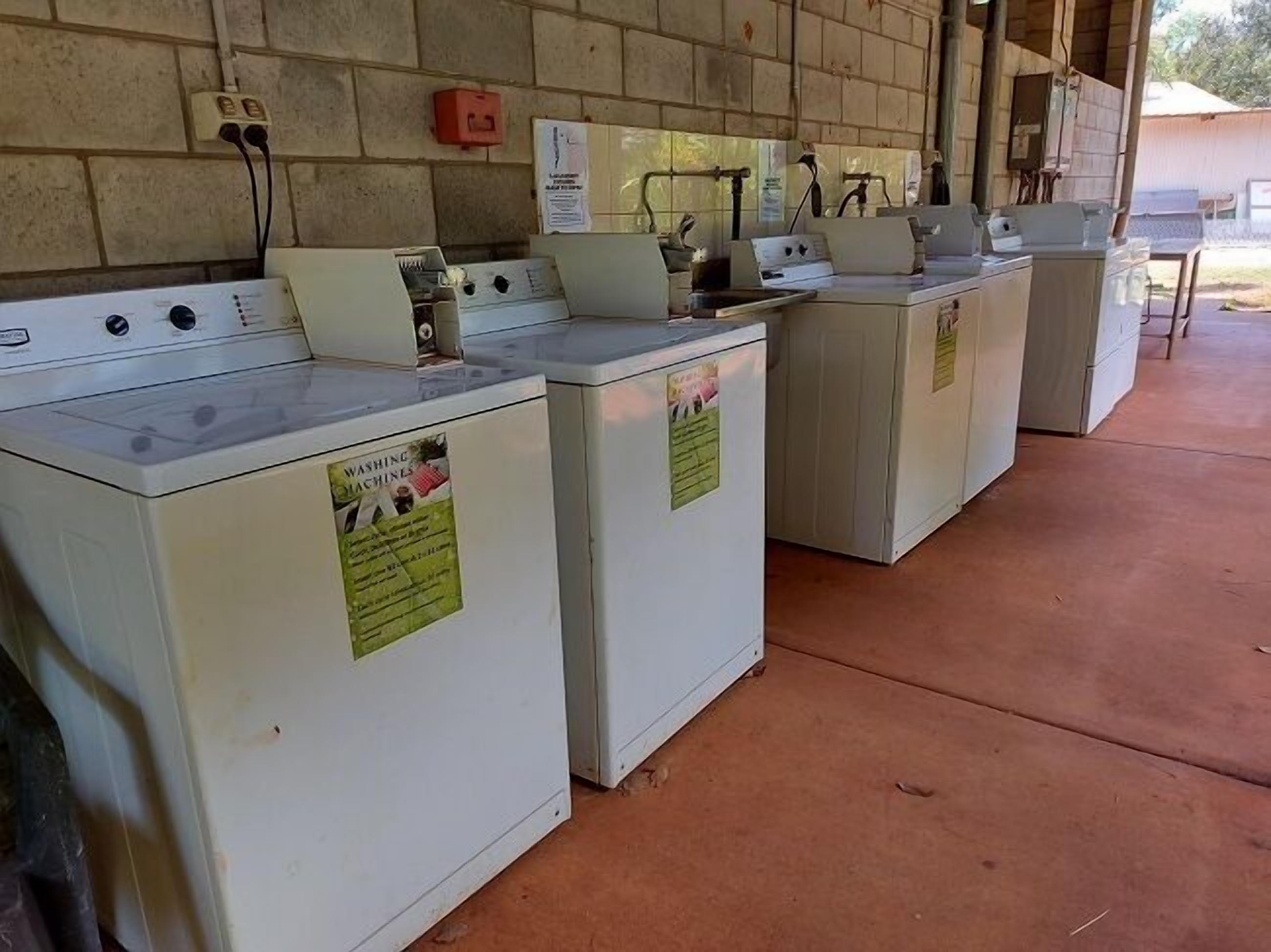 A row of white washing machines under a covered area with a tiled wall and electrical outlets.