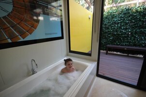 A bathtub with a child inside, a window with a yellow wall, and a wooden deck outside.