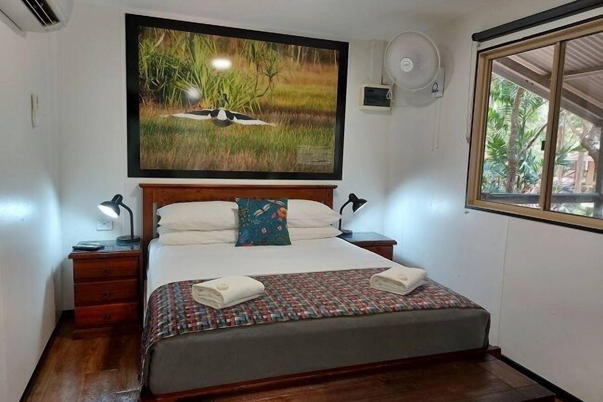 A bedroom with a bed, bedside tables, a wall-mounted air conditioner, a ceiling fan, and a window with a view of greenery.