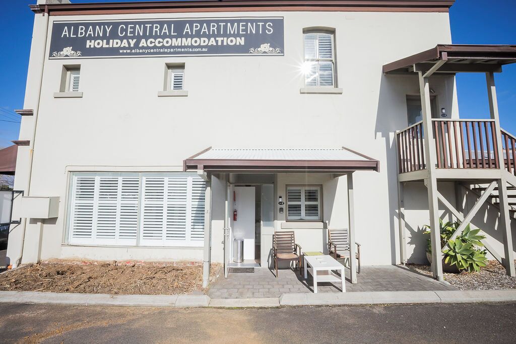 A two-story building with a sign for Albany Central Apartments Holiday Accommodation. There is a covered outdoor seating area with chairs and a table.