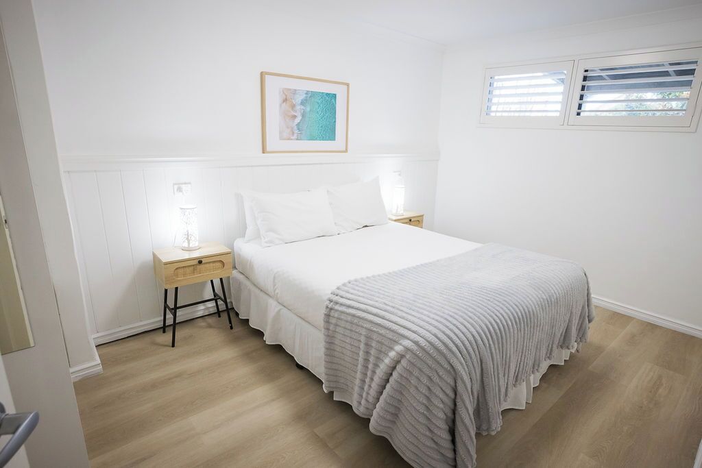Un dormitorio con cama, mesitas de noche, una obra de arte en la pared y una ventana con persianas.