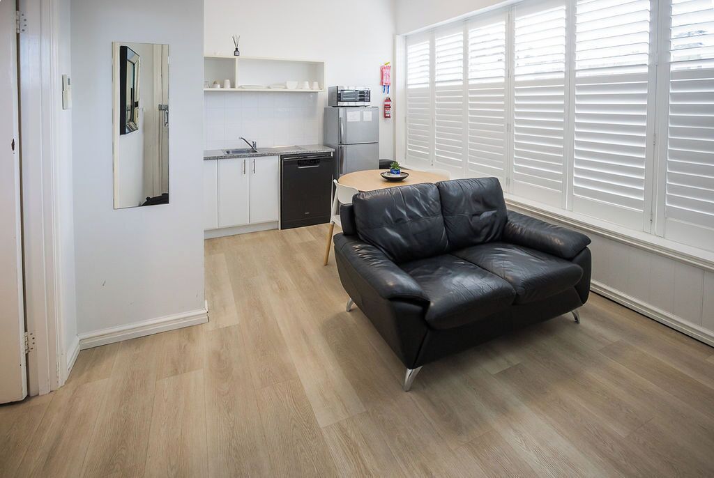 Una sala de estar moderna con un sofá de cuero negro, una mesa de comedor con sillas, una zona de cocina con armarios blancos y un ventanal con persianas.