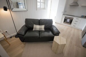 Una sala moderna con un sofá de cuero negro, un tapete con patrón de espiga y una cocina con muebles blancos y horno empotrado.