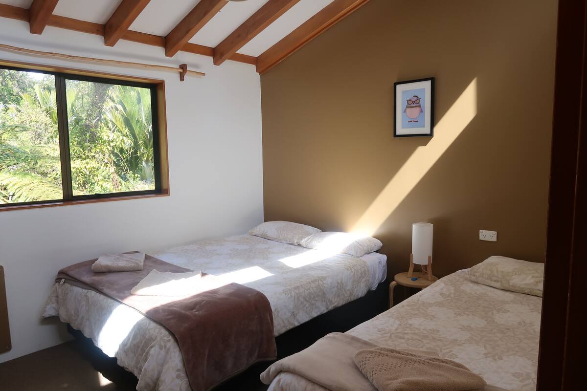 A room with two single beds, a window with a view of greenery, a framed picture on the wall, and a bedside table with a lamp.