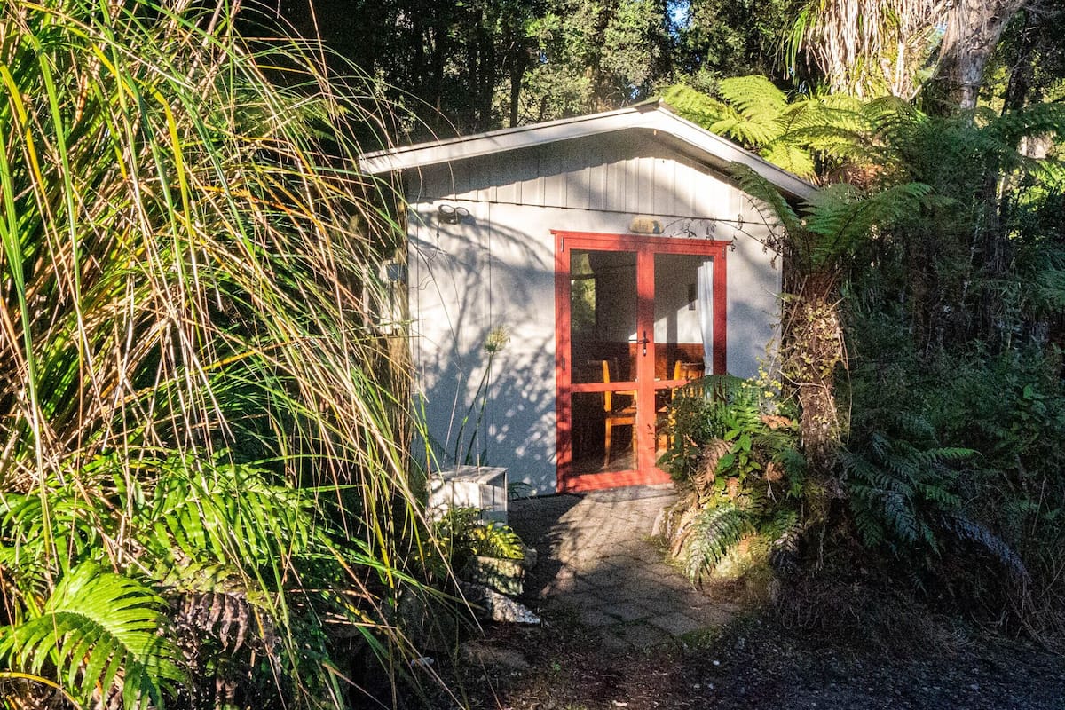 A small, single-story building with a red door and a window, surrounded by dense greenery and trees.