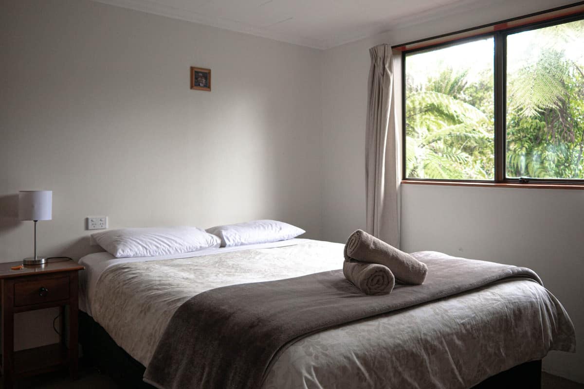 A bedroom with a bed, a nightstand, a lamp, and a window with a view of greenery.