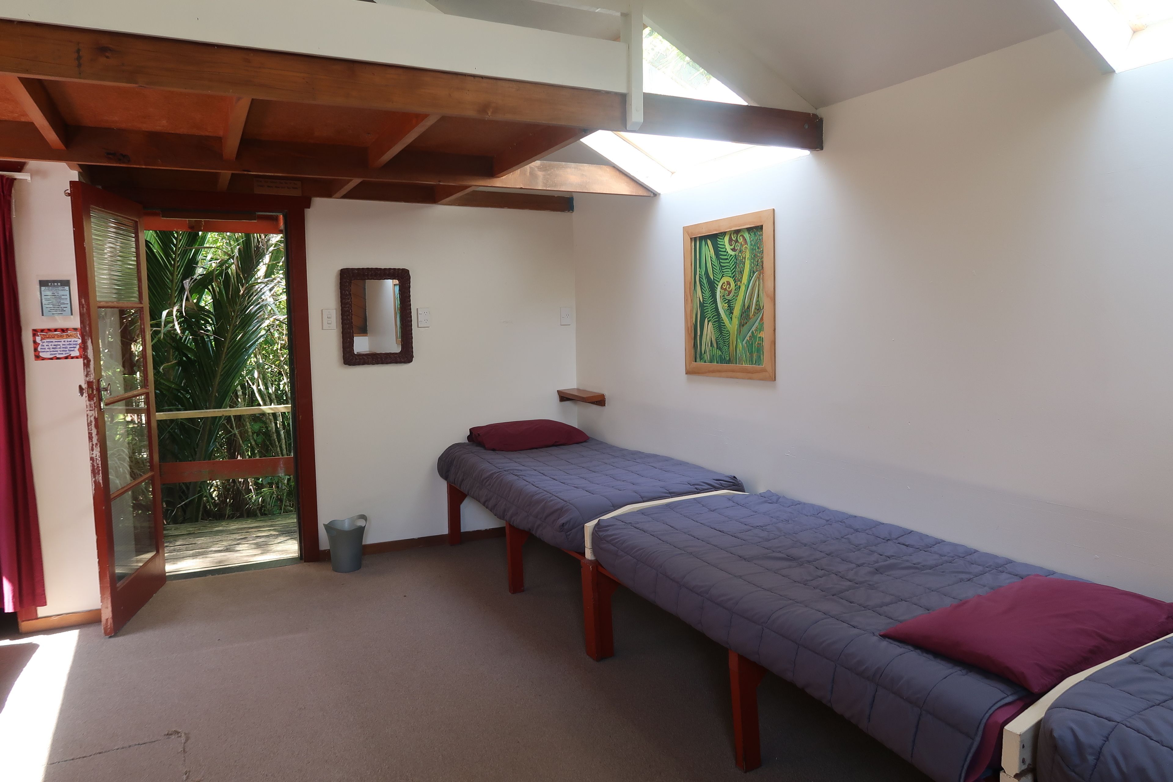 A room with two single beds, a wooden ceiling, a small window, and a door leading outside.