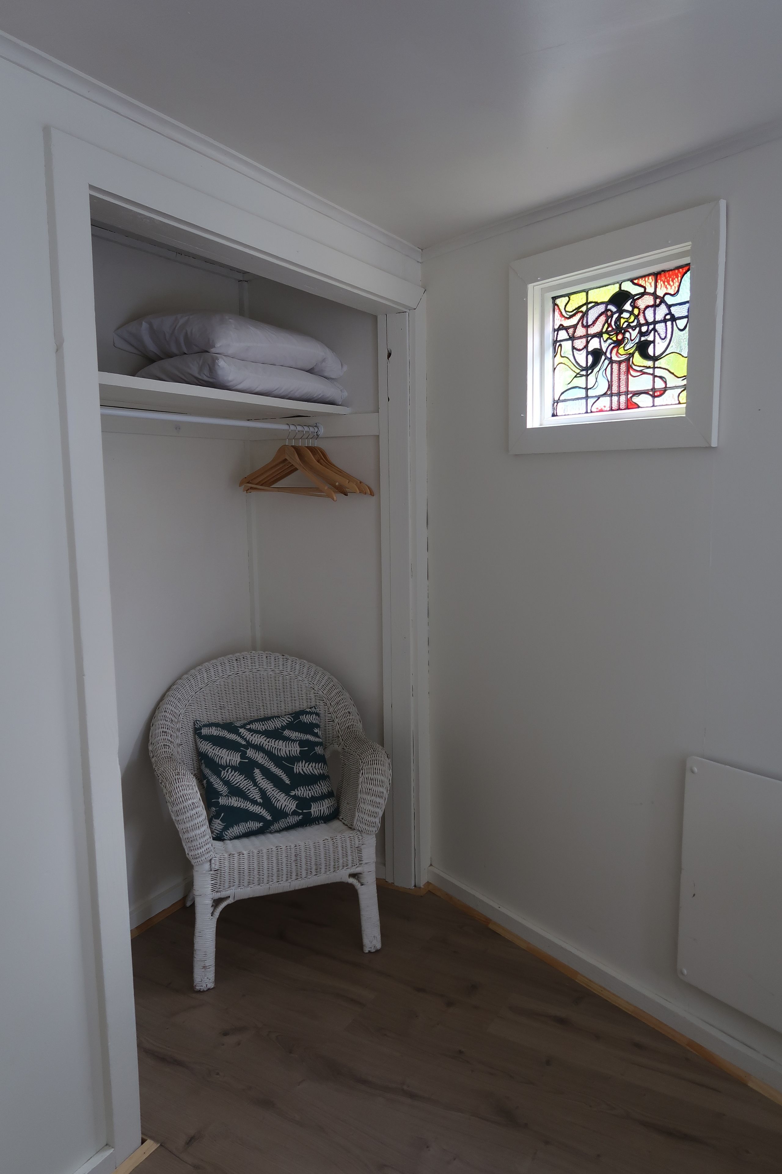 A white room with a wicker chair, a hanging clothes rack, and a window with stained glass.