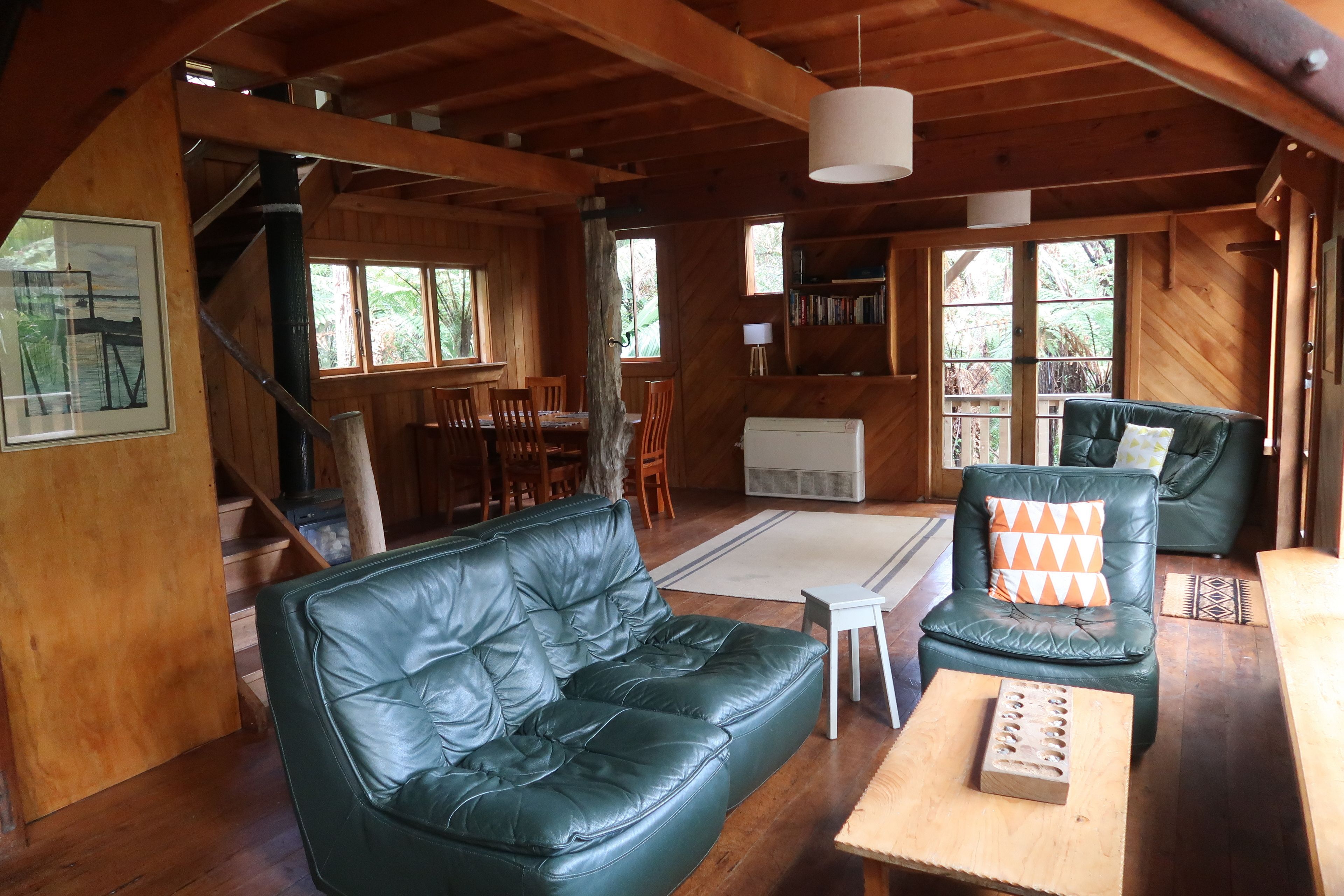 Un interior de cabaña acogedor, con una escalera de madera, un sofá de cuero verde, una zona de comedor y una estantería.