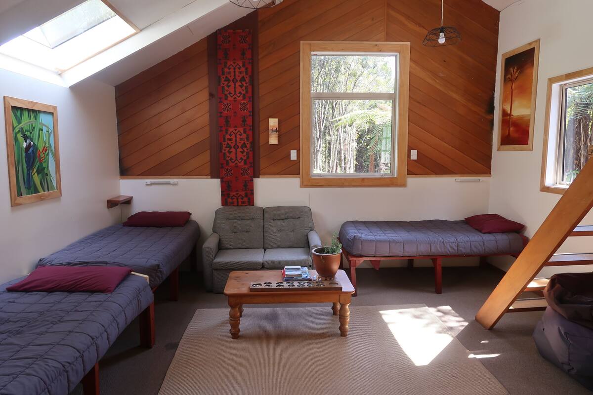 A cozy room with two single beds, a sofa, a wooden table with a plant, and a large window with a view of trees.