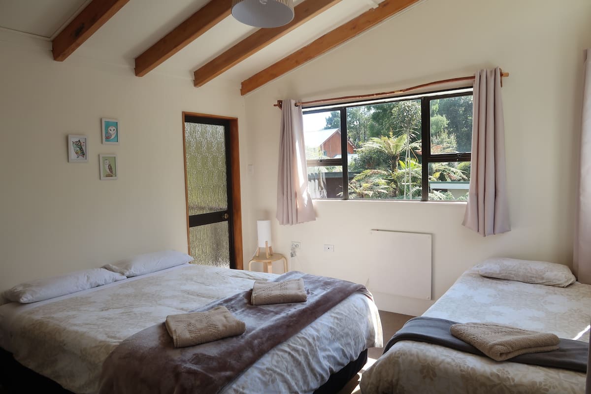 A room with two beds, a window with curtains, a radiator, and a small table with a lamp.