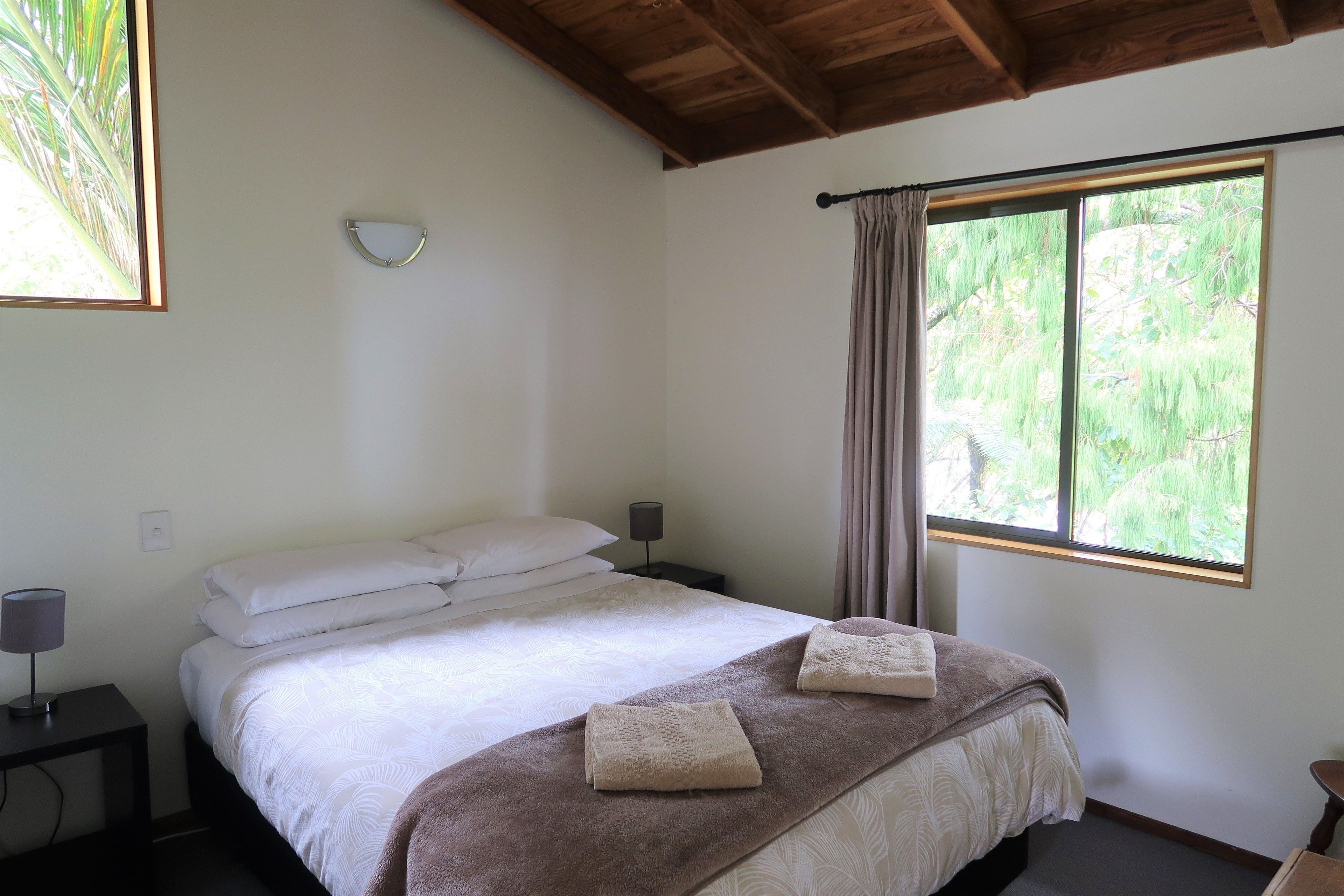 Un dormitorio con una cama, dos mesitas de noche, una mesita auxiliar, una ventana con cortinas y vistas a la vegetación.
