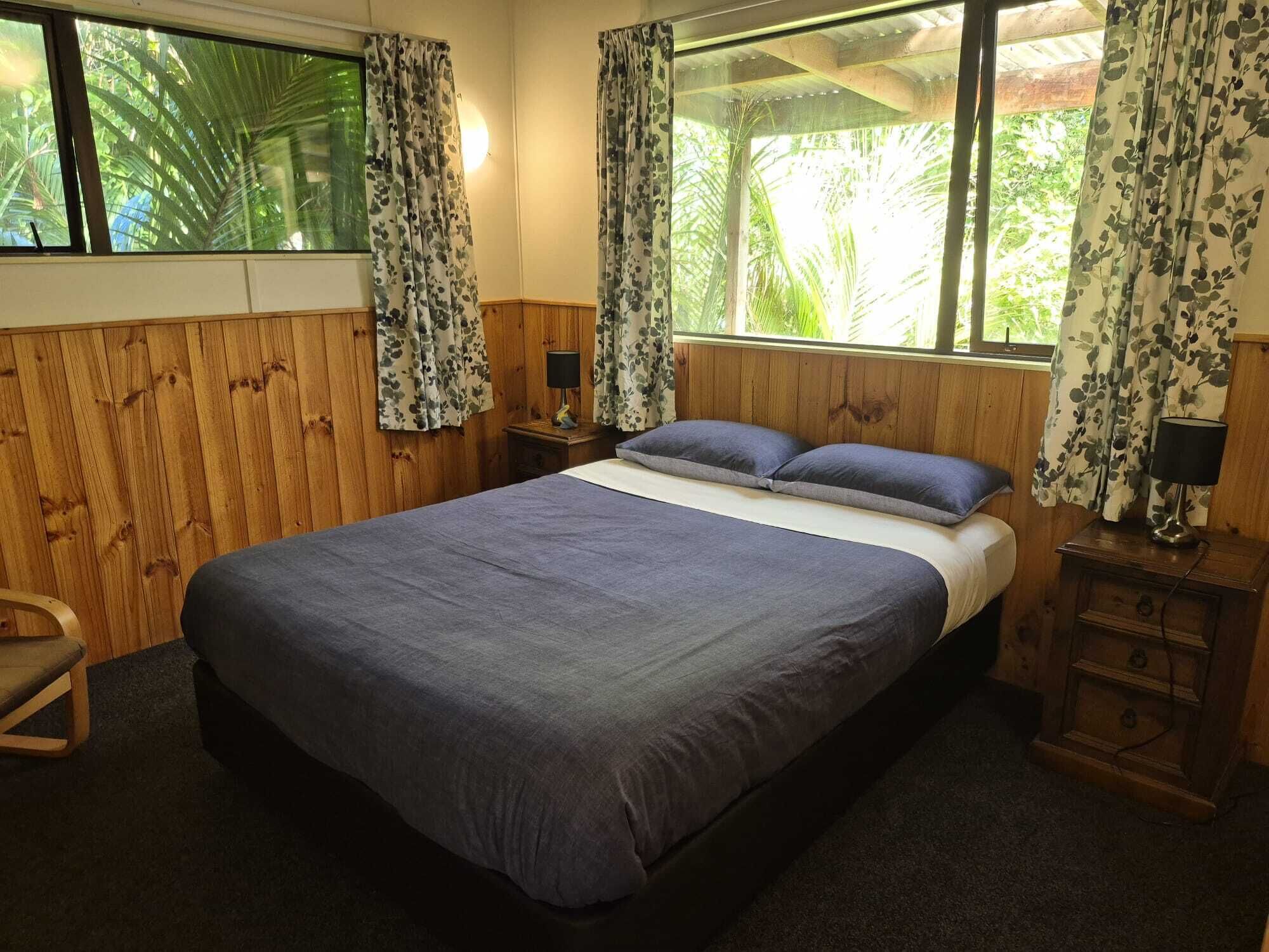 A room with a large bed, wooden walls, a window with curtains, a nightstand with a lamp, and a chair.