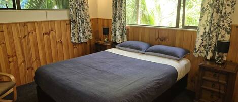 A room with a large bed, wooden walls, a window with curtains, a nightstand with a lamp, and a chair.