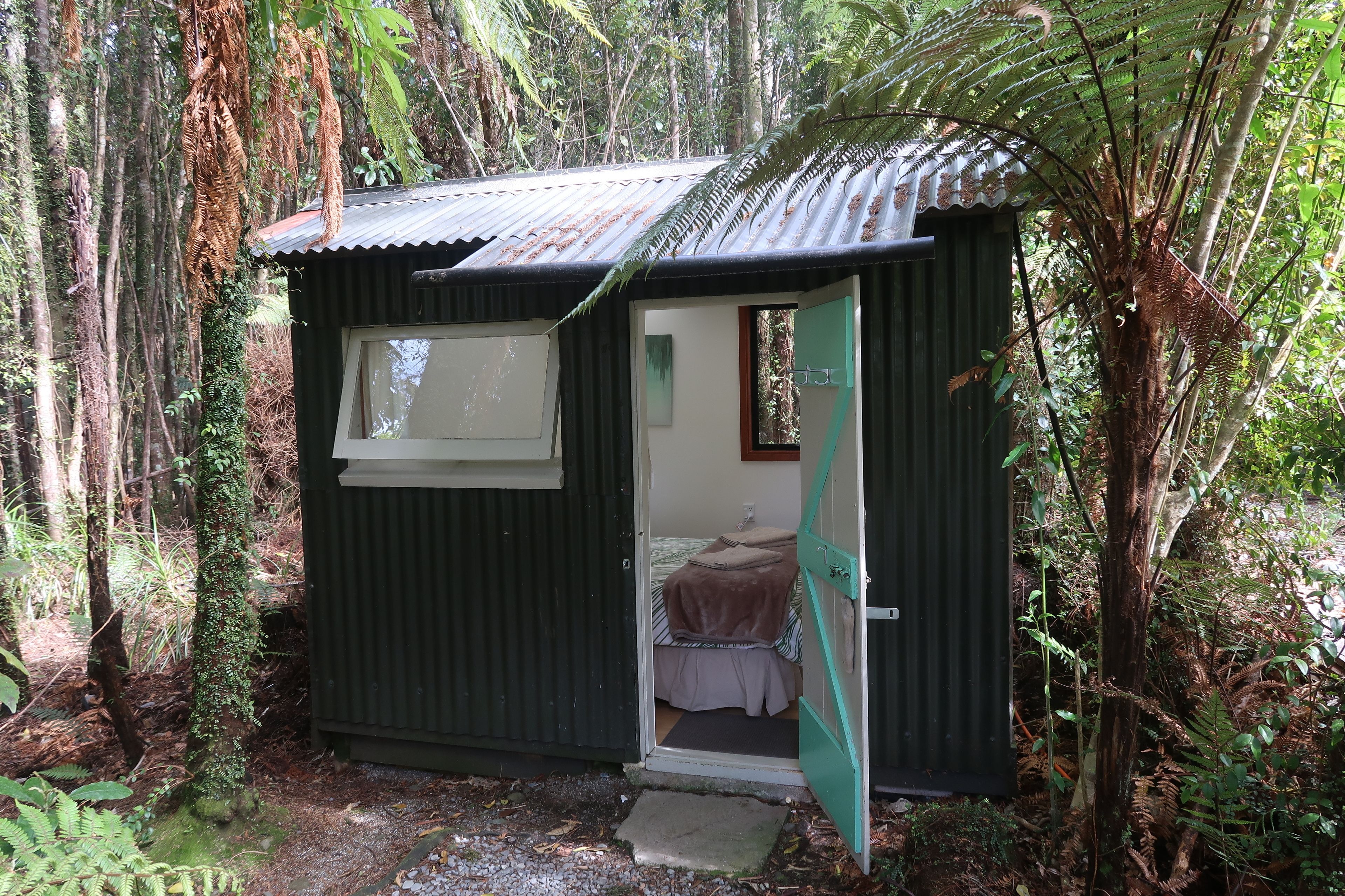 A small, rustic cabin with a single bed and a window, surrounded by dense forest.