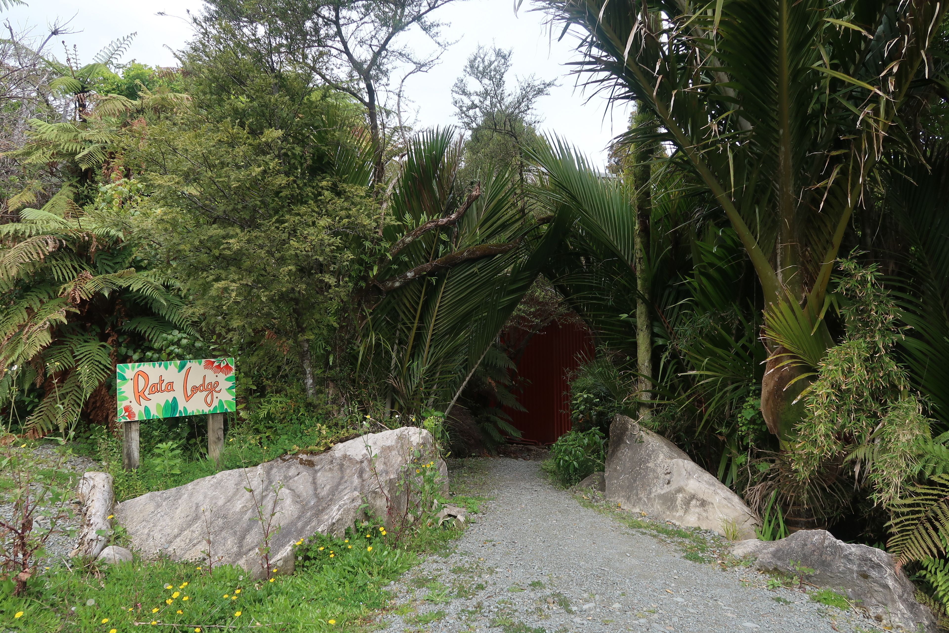 Ein Weg führt zu einer roten Tür, umgeben von üppigem Grün und einem Schild mit der Aufschrift 'Rata Lodge'.