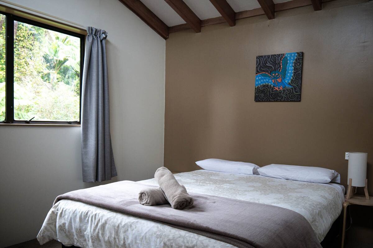 A bedroom with a bed, a window with curtains, a painting on the wall, and a bedside lamp.