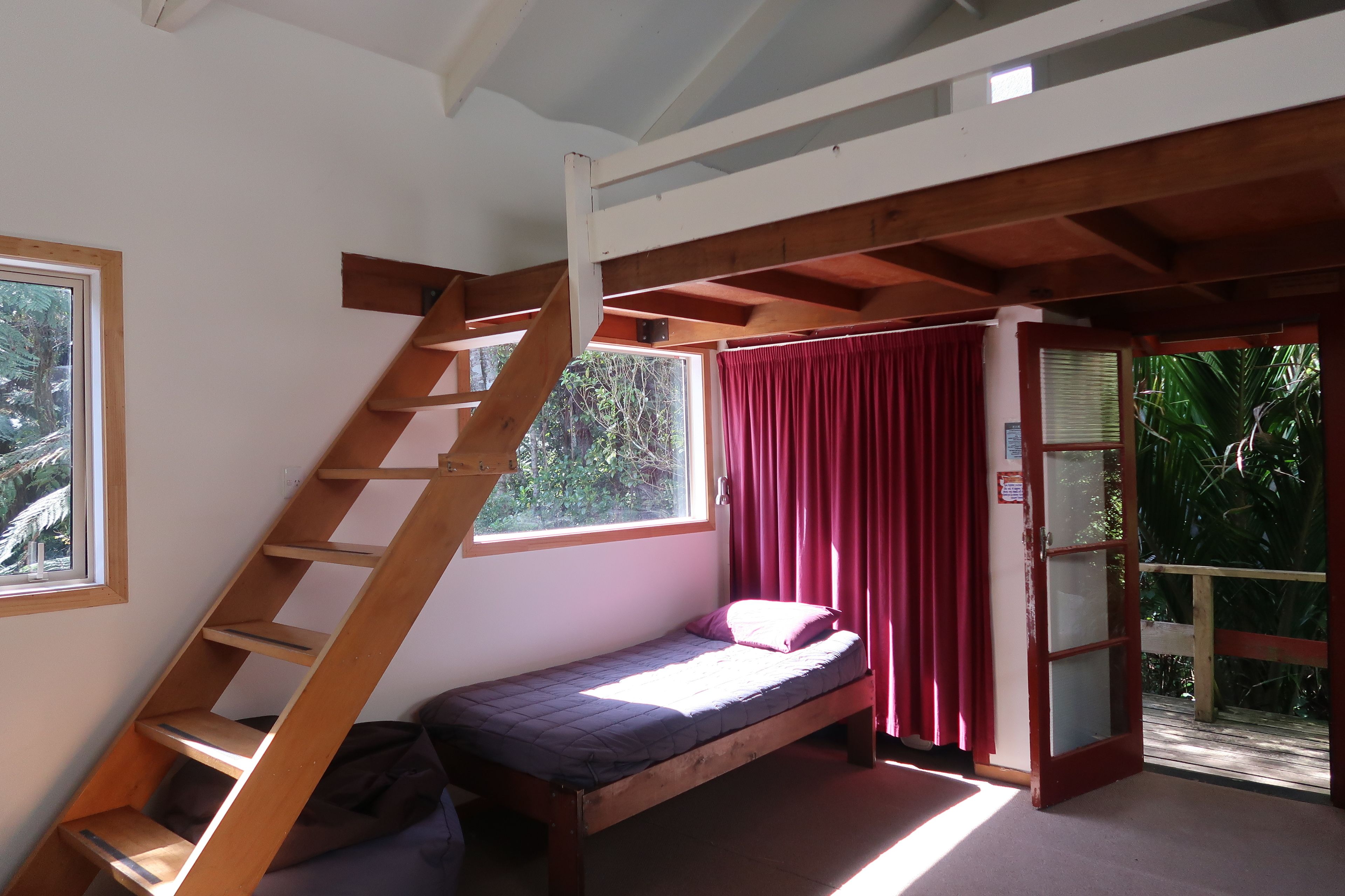 Una habitación pequeña con cama individual, una escalera de madera que lleva a una zona superior, una ventana con cortinas y una puerta que da a un espacio exterior.