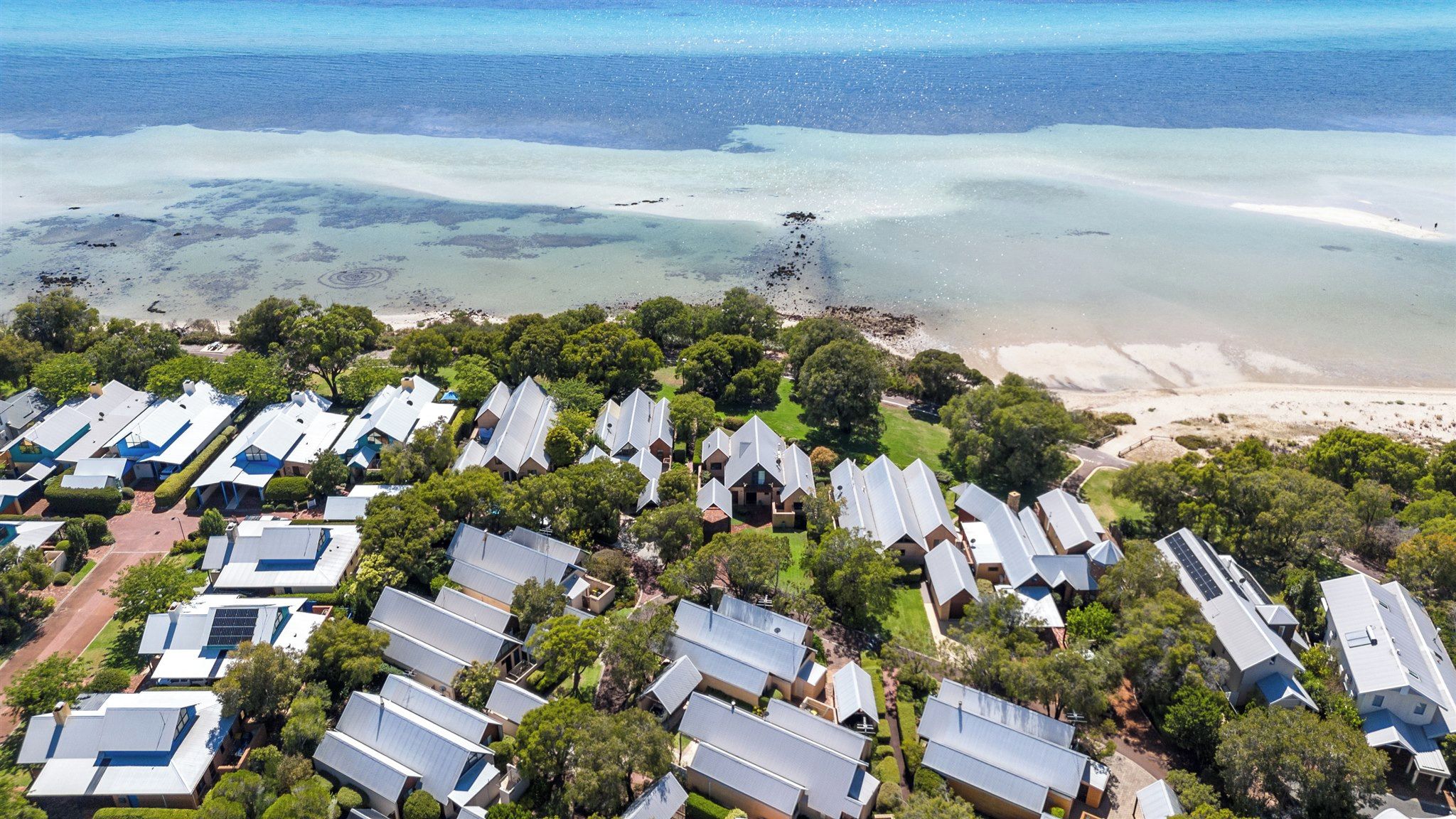 A coastal residential area with multiple houses featuring sloped roofs, likely with solar panels, surrounded by greenery and a beach.