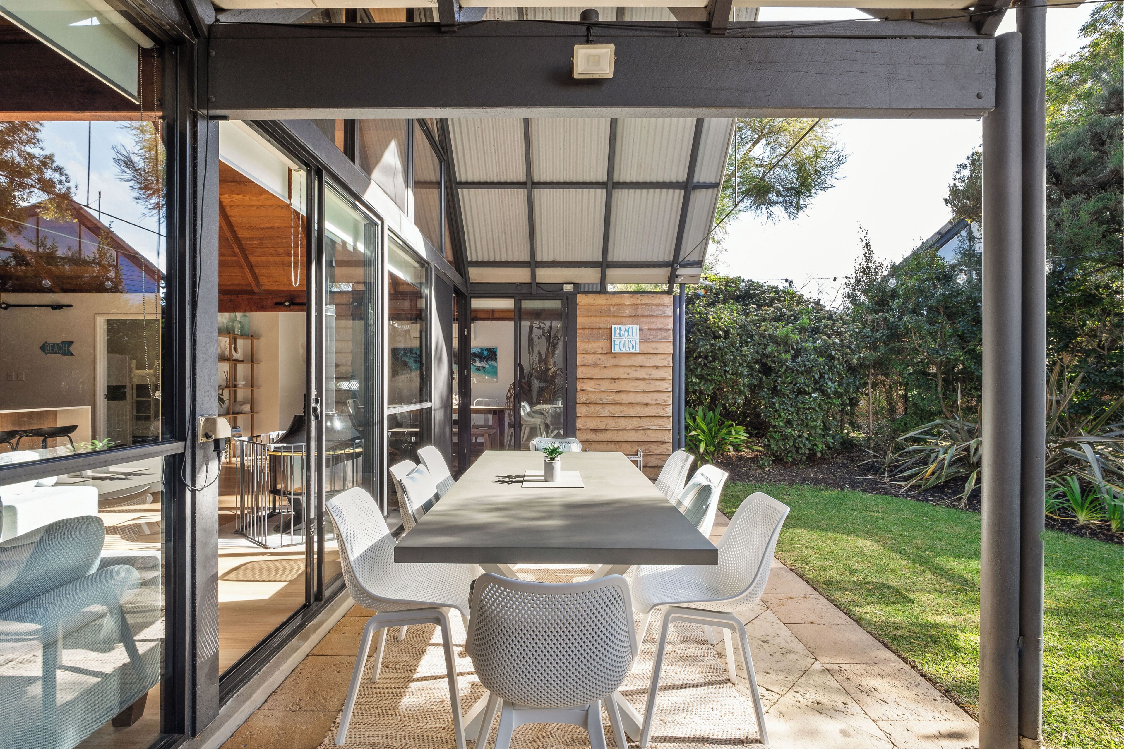 An outdoor dining area with a long table and white chairs, a covered patio, and a view of the house interior.