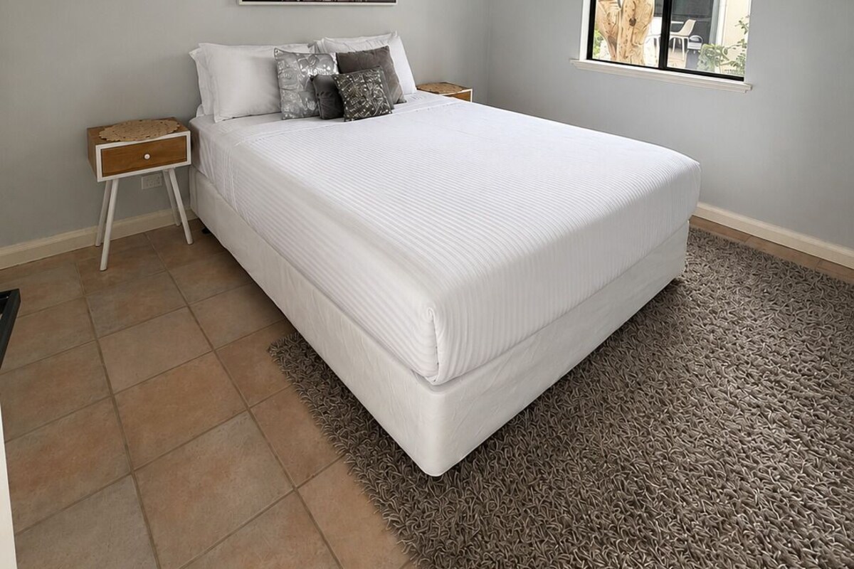 A bedroom with a bed, a nightstand, a wall-mounted lamp, a window, and a ceiling light fixture.
