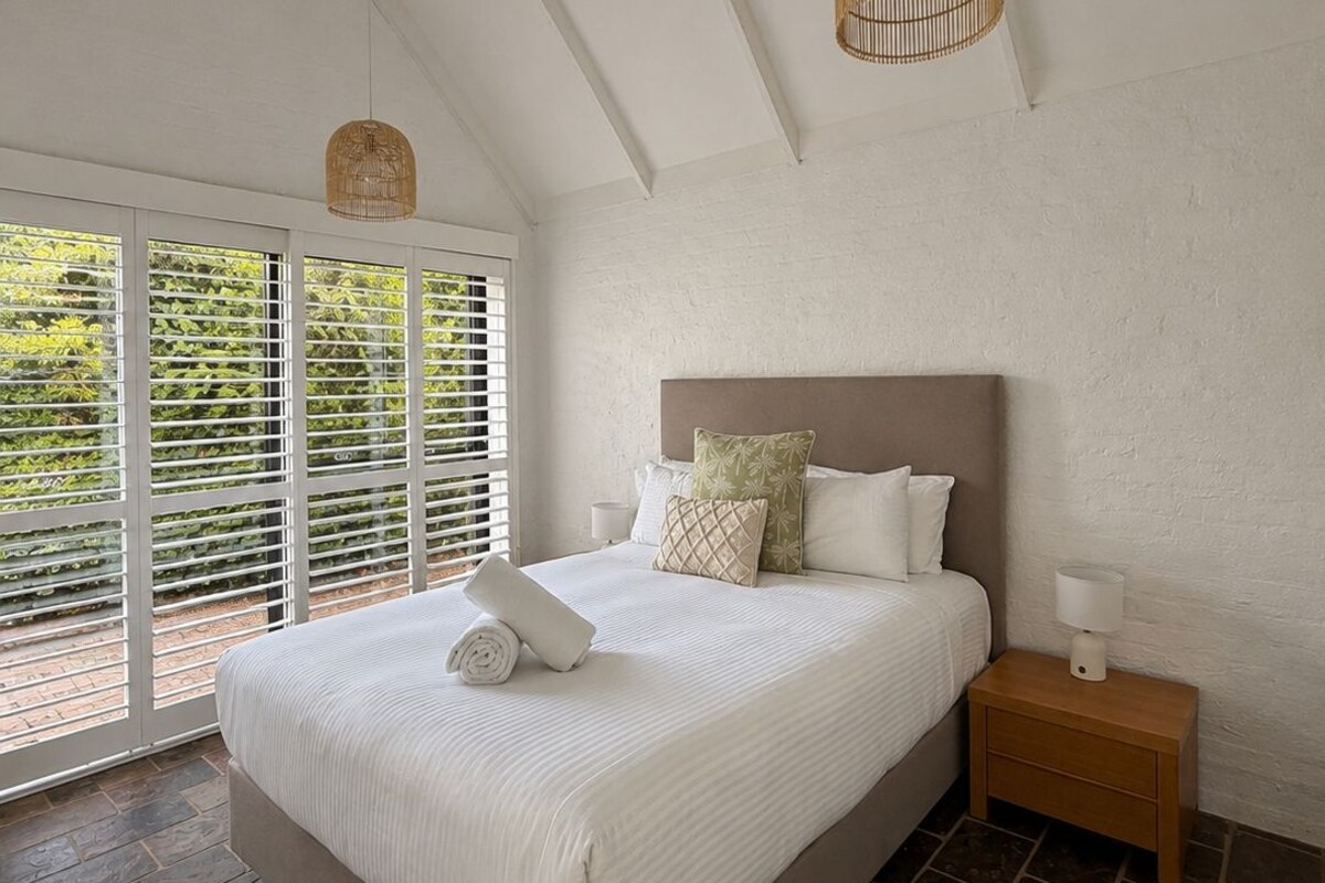 A bedroom with a large bed, a ceiling fan, a nightstand, and a window with blinds.